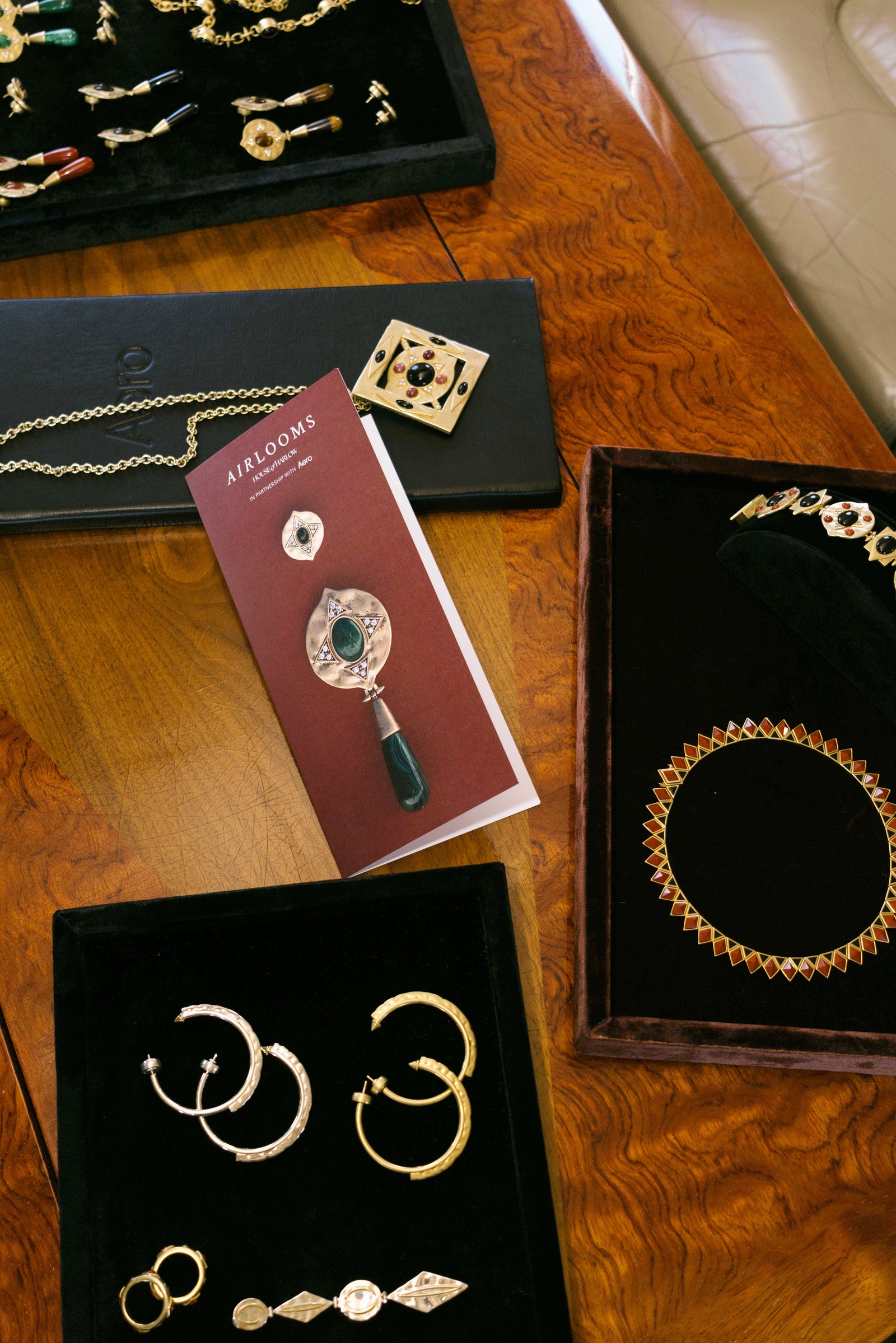 An AIRLOOMS brochure laid out with House of Harlow chain necklaces, gold hoop earrings, a collar necklace and jewelry trays on a burl wood table.