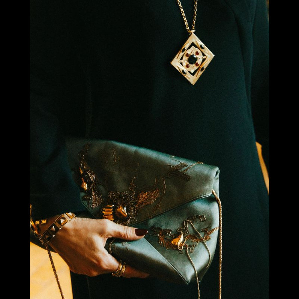 The Hathor Spinning Pendant Necklace in black onyx worn over a dark green dress, with a hand holding an embroidered clutch.
