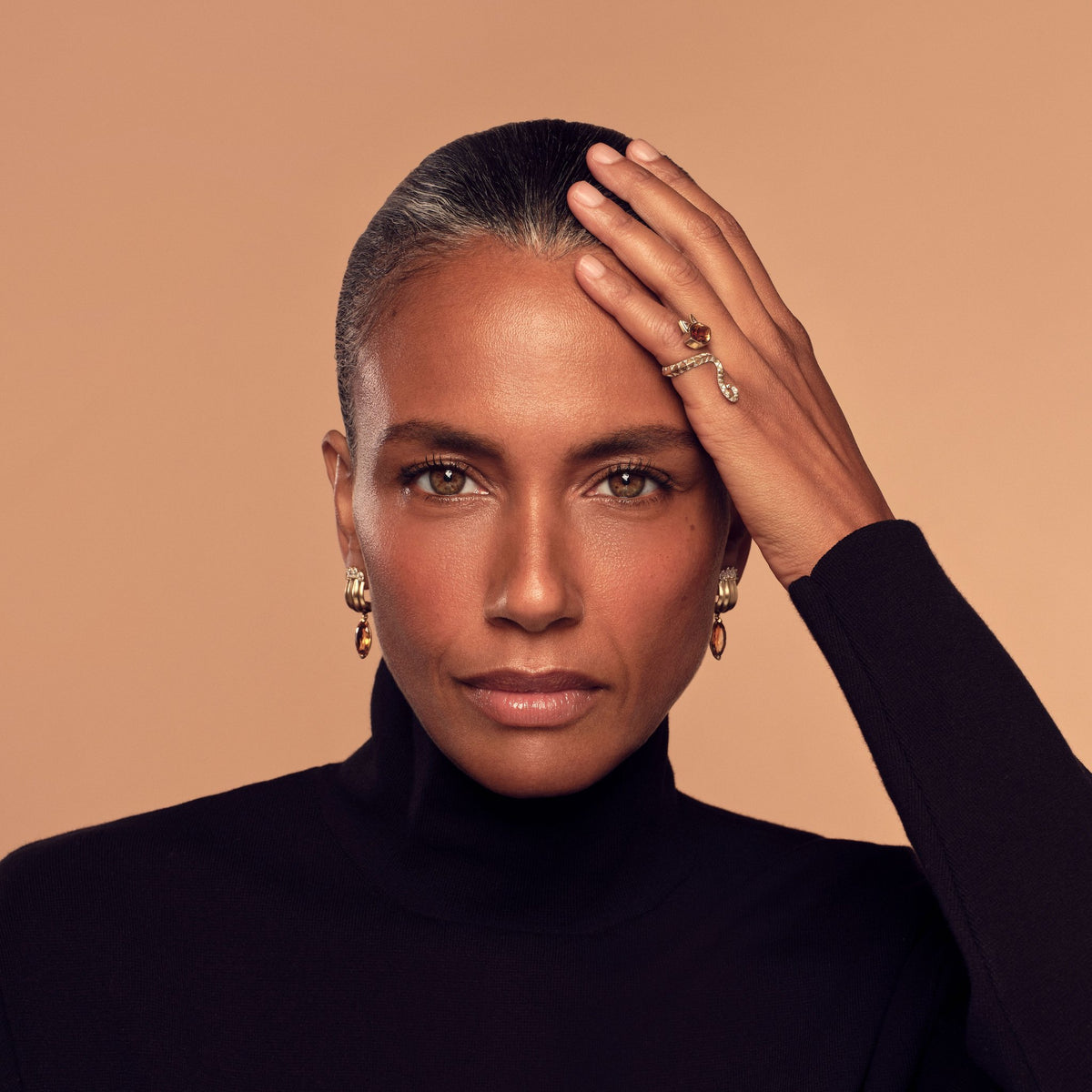 Model in a black turtleneck with a hand to her forehead, wearing House of Harlow gold rings and earrings against a peach backdrop.