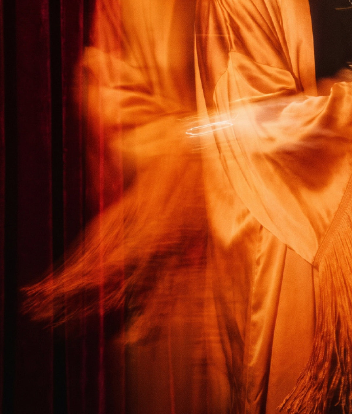 A figure in a flowing orange veil blurred in motion against a dark red curtain.