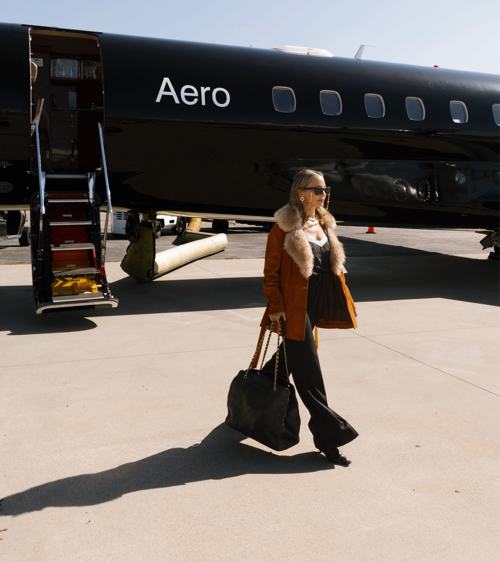 A traveller in a rust shearling coat and House of Harlow fine jewelry walks across the tarmac to a waiting Aero private jet.