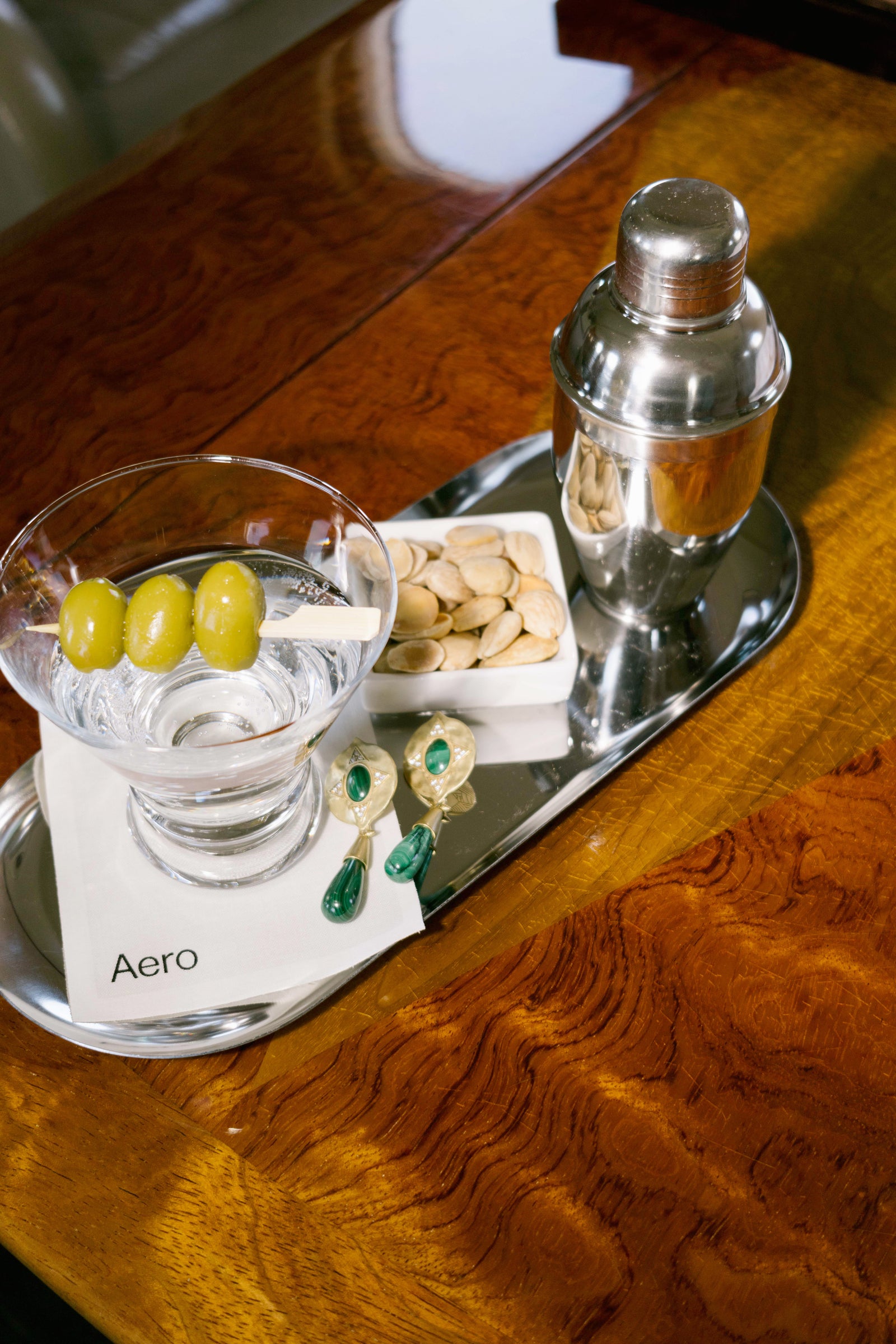 House of Harlow Hathor Teardrop Earrings in malachite resting on an Aero tray beside a martini, cocktail shaker and pistachios on a burl wood table.