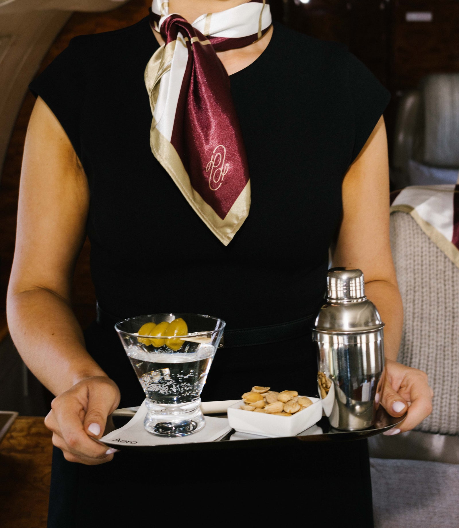 An Aero flight attendant in a printed silk scarf carries a tray with a martini, cocktail shaker and almonds during the AIRLOOMS in-flight experience.