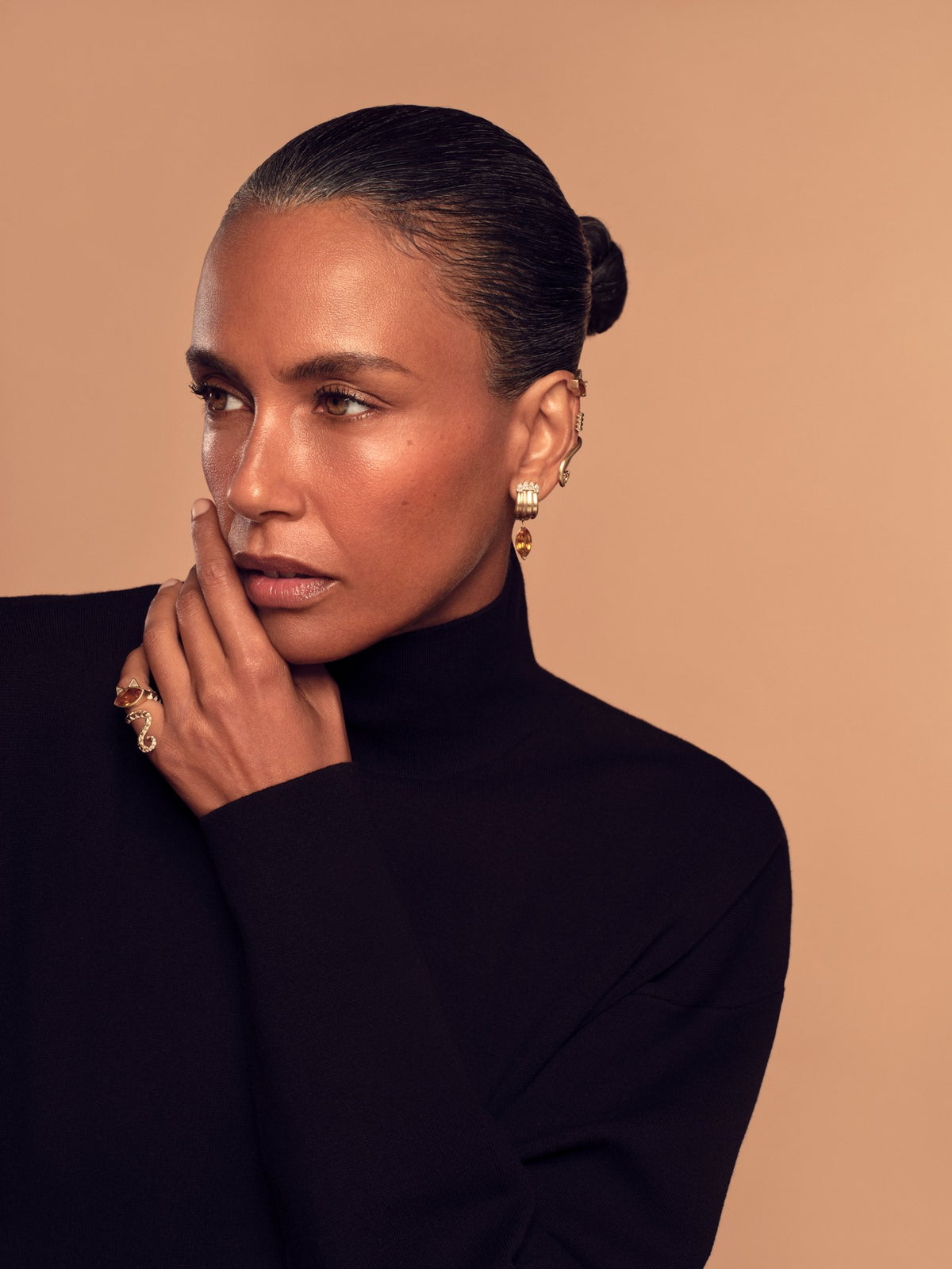 Model in a black turtleneck with a hand at her chin, wearing House of Harlow gold earrings and rings against a peach backdrop.