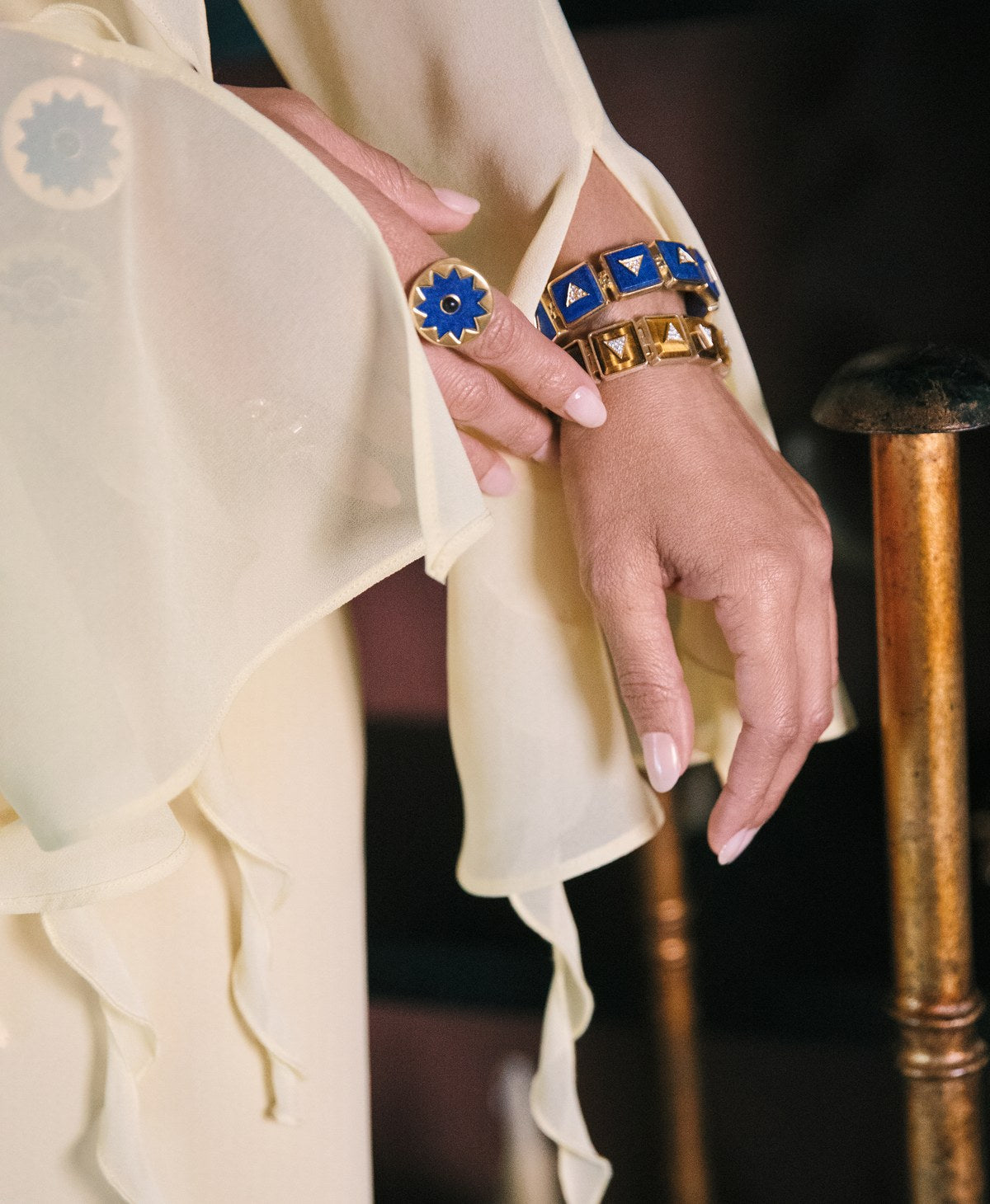 Hand resting on a gilded chair wearing the Sunburst Statement Bracelet and Sunburst Cocktail Ring in blue lapis